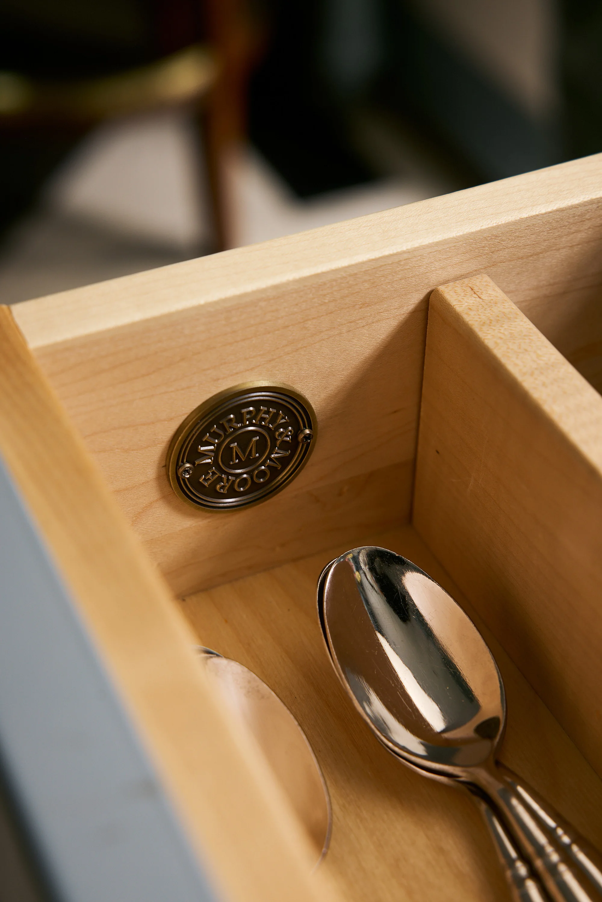 Interior view of a light-wood drawer with silverware spoons inside and a round, bronze Murphy & Moore logo affixed to the side.