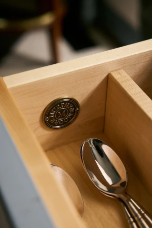 Interior view of a light-wood drawer with silverware spoons inside and a round, bronze Murphy & Moore logo affixed to the side.