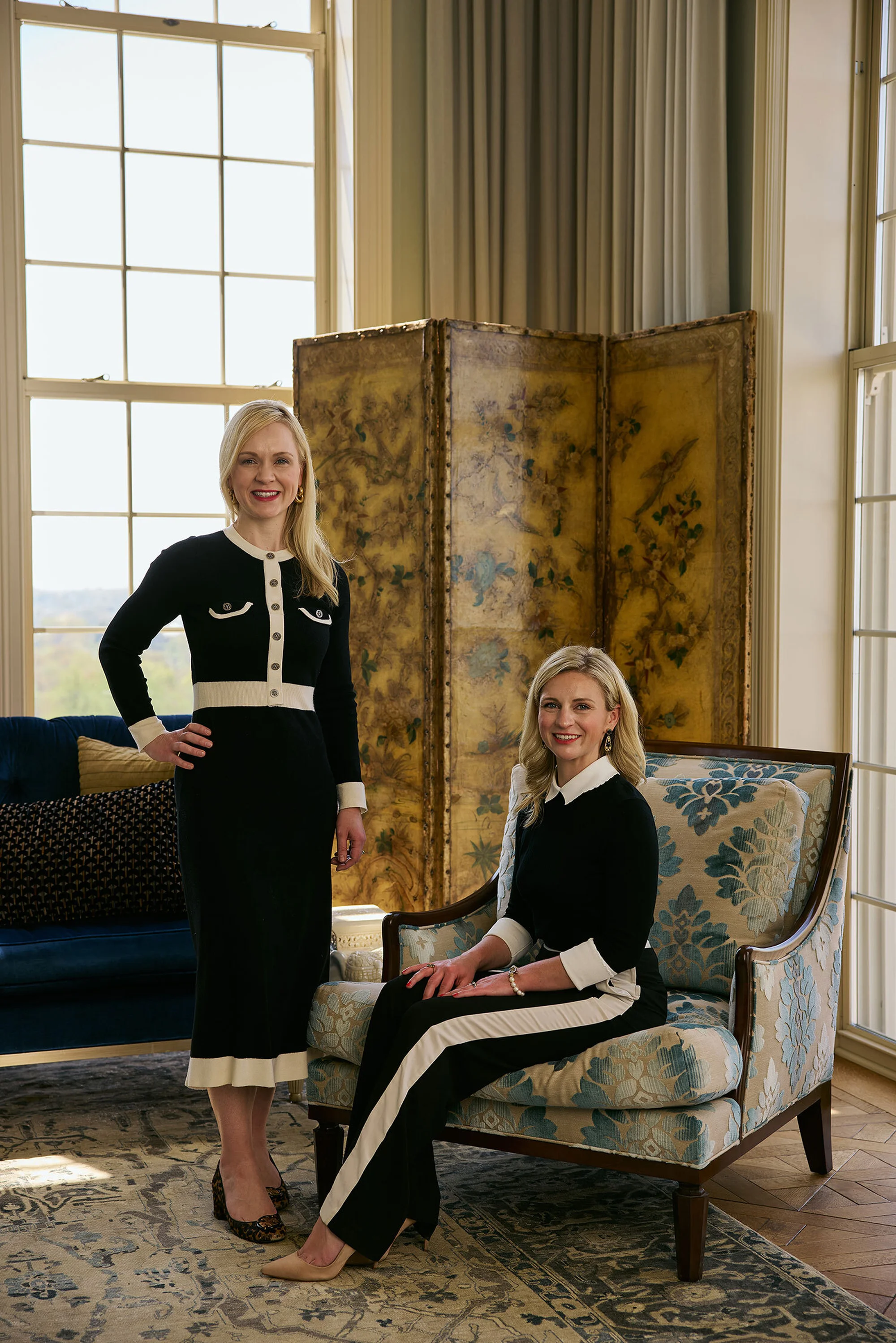 Two fair-skinned adult women pose indoors in black and white outfits. One woman sits in a patterned chair, the other stands next to a sofa.