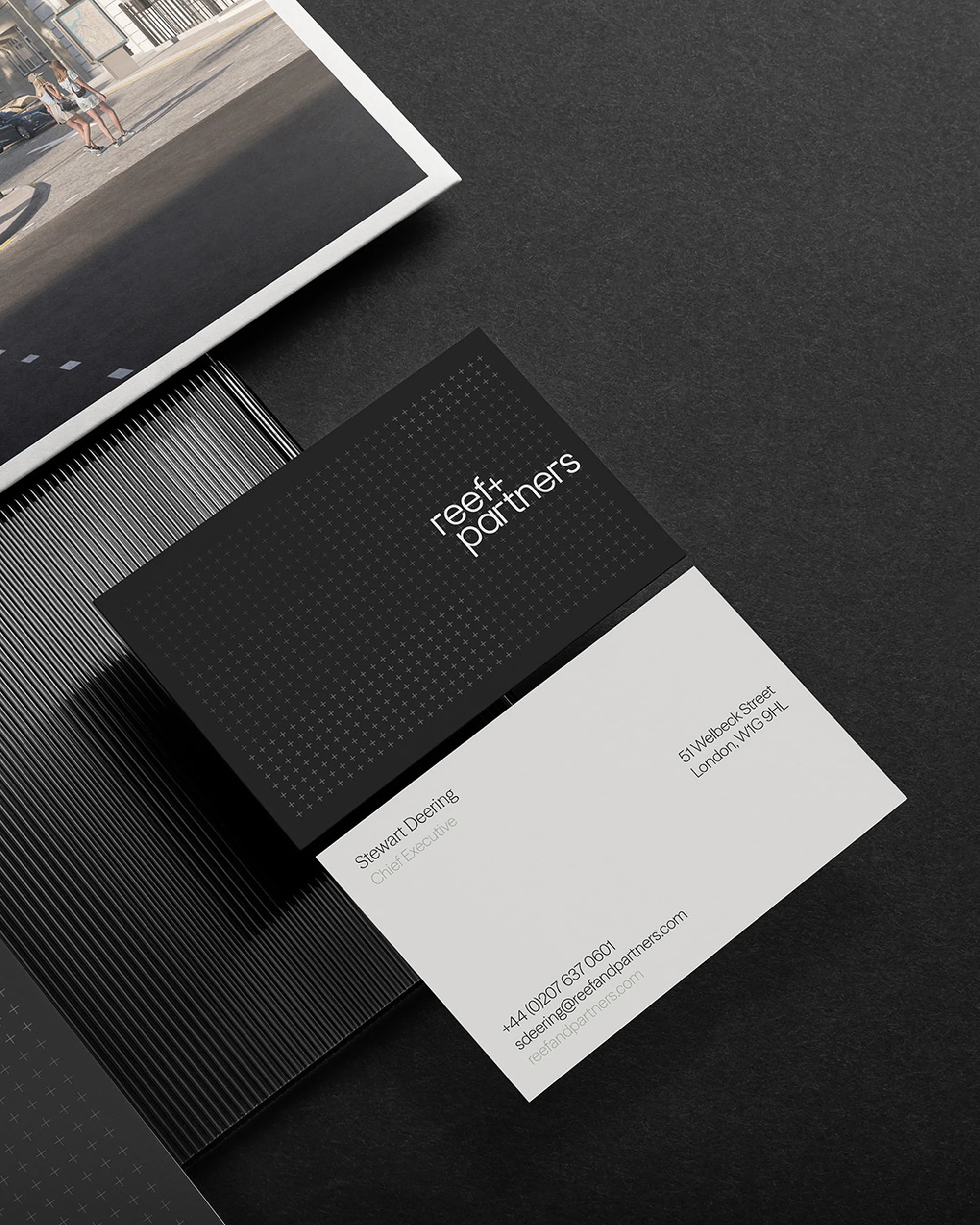 Black and white business cards with the name 'reef+partners' and contact details, presented on a textured black surface, studio setting.
