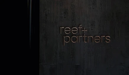 Bronzed 3D typography of Reef plus Partners mounted on a dark charred wood panel wall.