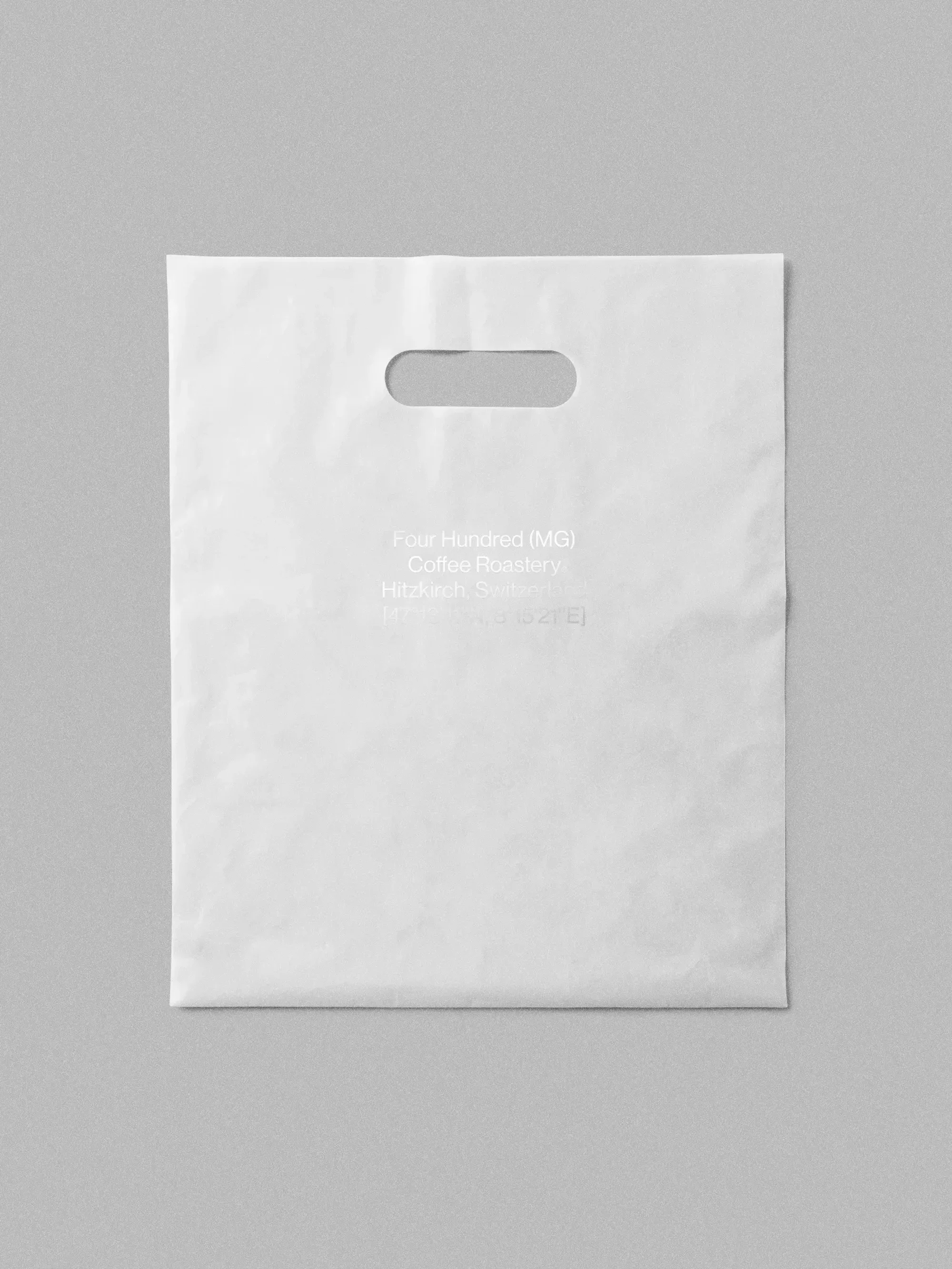 Isolated white plastic shopping bag with cutout handles on a gray surface, featuring a coffee roastery logo and branding text.