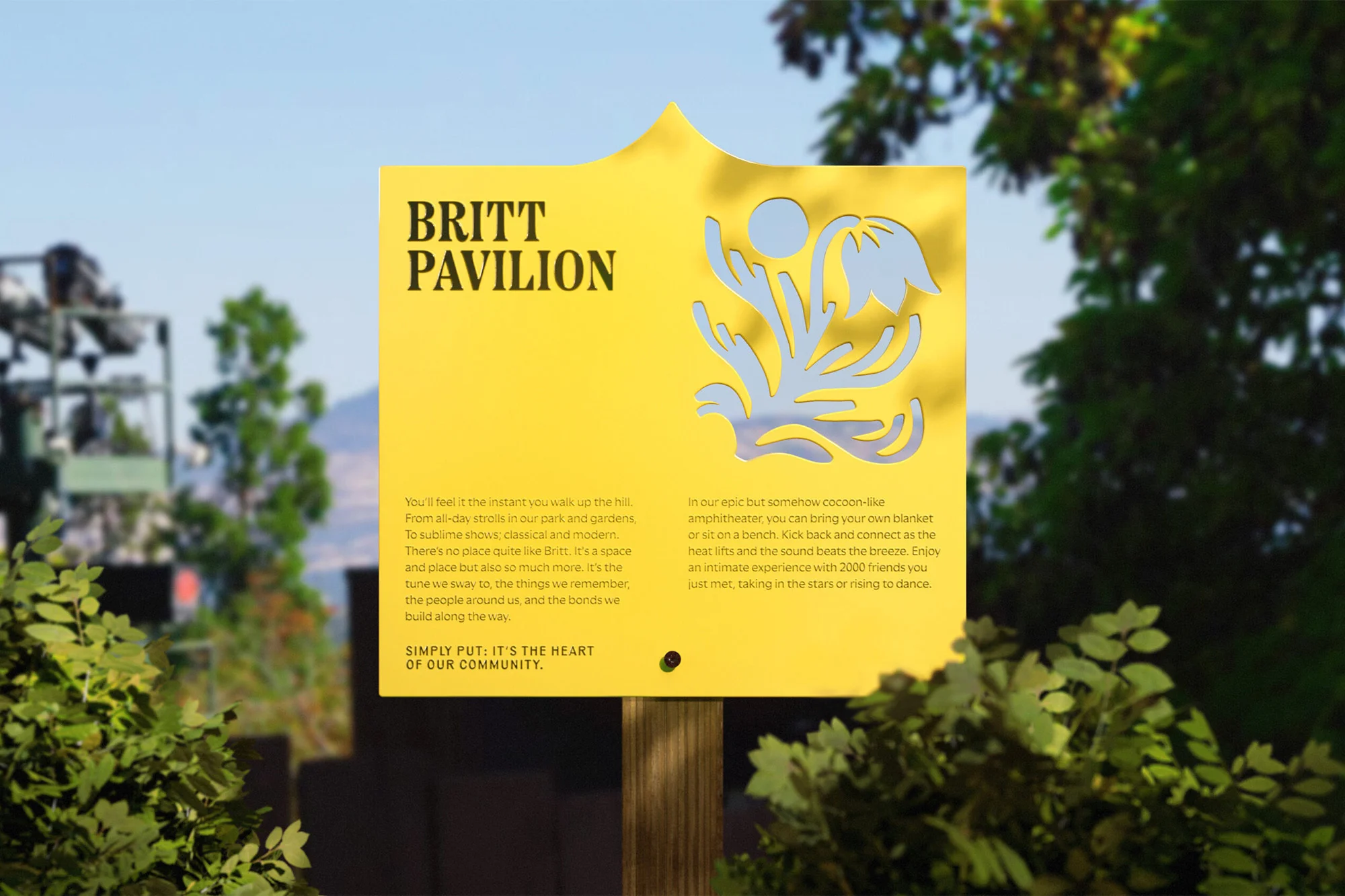 Yellow sign for Britt Pavilion in a park setting. Sign features a stylized flower graphic. Trees and sky in the background.