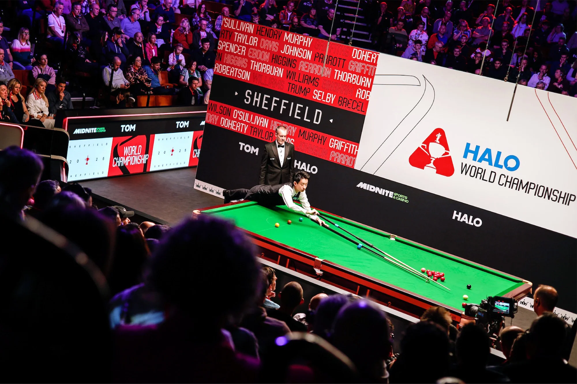 Snooker match at the Halo World Championship in Sheffield: a player lines up a shot, a referee watches, and spectators observe from the stands.