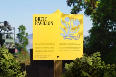 Bright yellow information sign featuring black typography reading Britt Pavilion and a cutout graphic set outdoors.