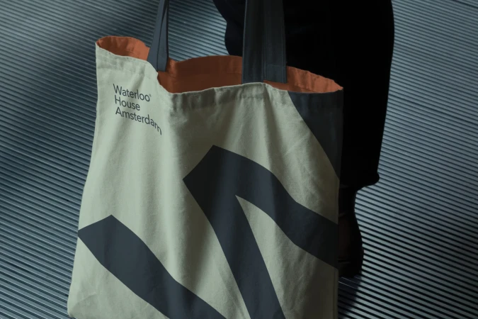 Tan tote bag with grey graphic and handle, featuring 'Waterloo House Amsterdam' text, is carried against a grey ribbed wall.