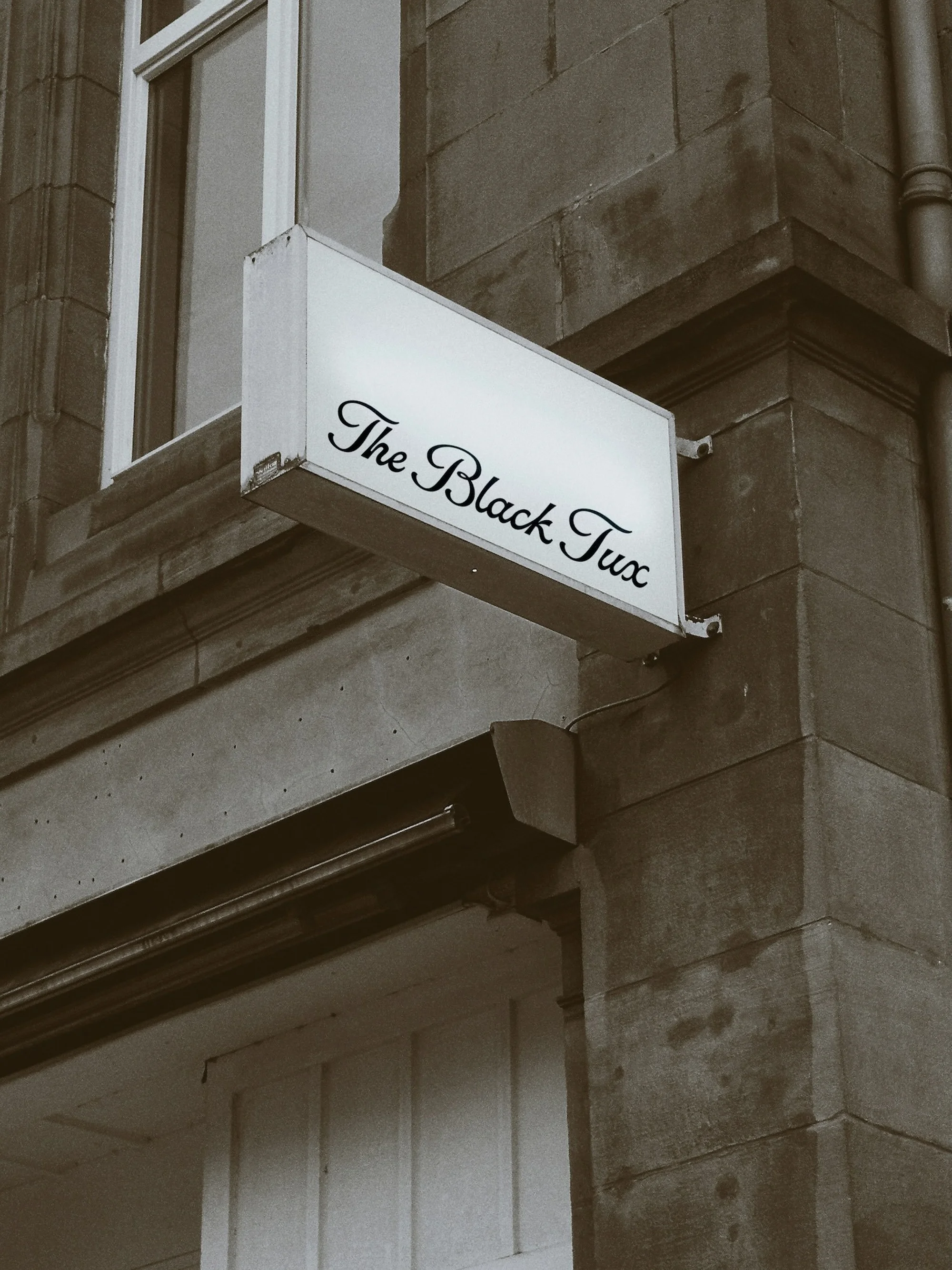 Illuminated sign reading 'The Black Tux' on the exterior of a stone building with a window above, under overcast lighting.