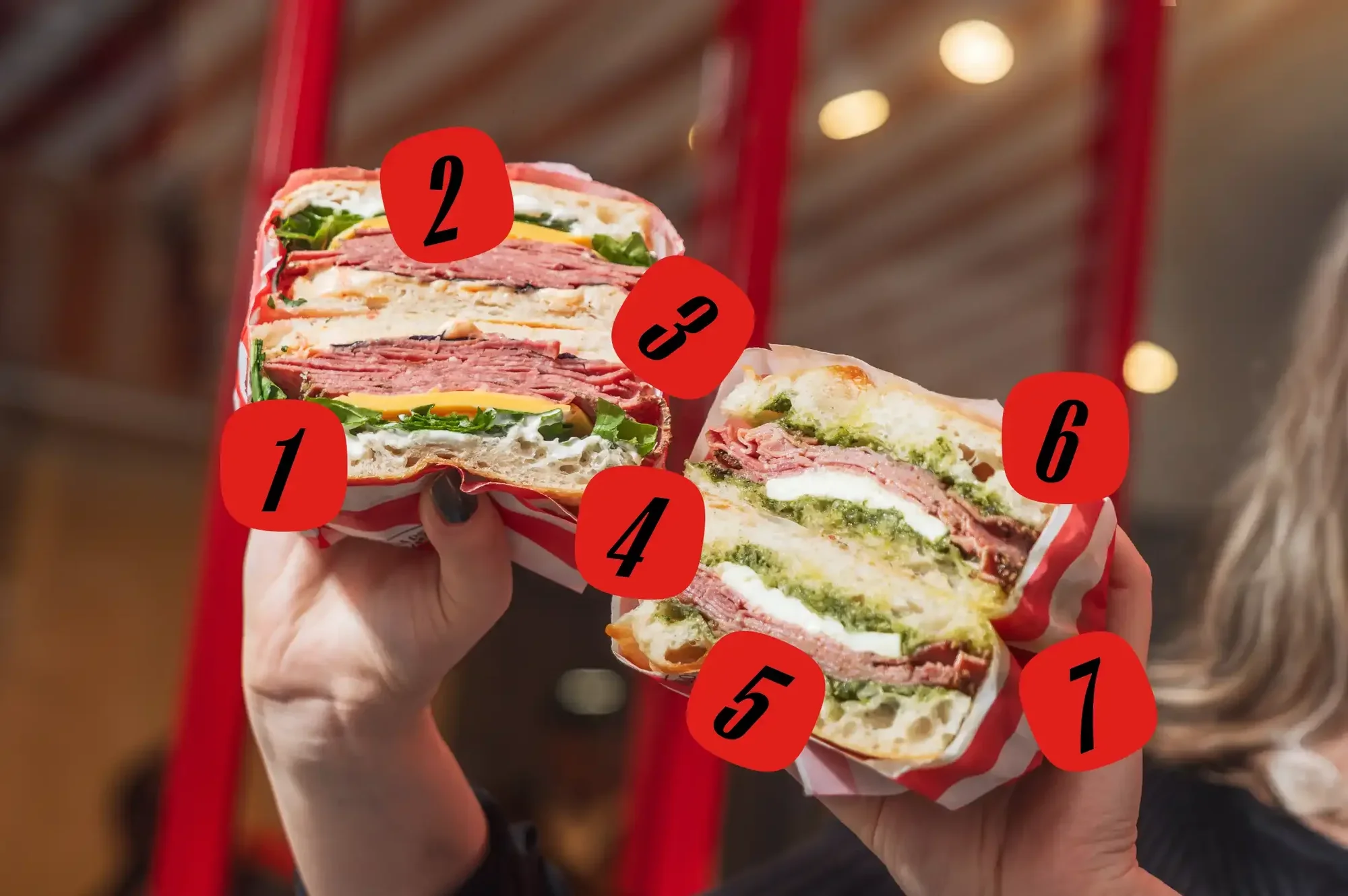 Two sandwich halves with lettuce, tomato, cheese, and meat are held by a person in front of a red and white striped background.