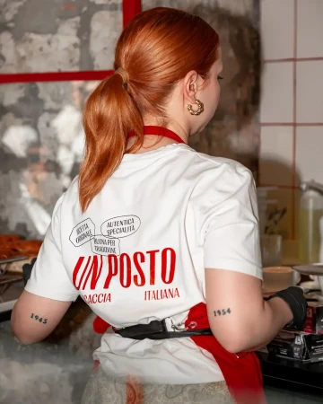 Woman with red hair in ponytail wearing a white t-shirt with red Italian text, standing in a kitchen-like environment.