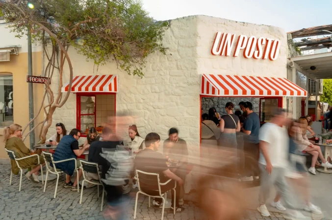 Outdoor cafe scene with people seated at tables and blurred pedestrians passing by the entrance of a brick building with striped awnings.