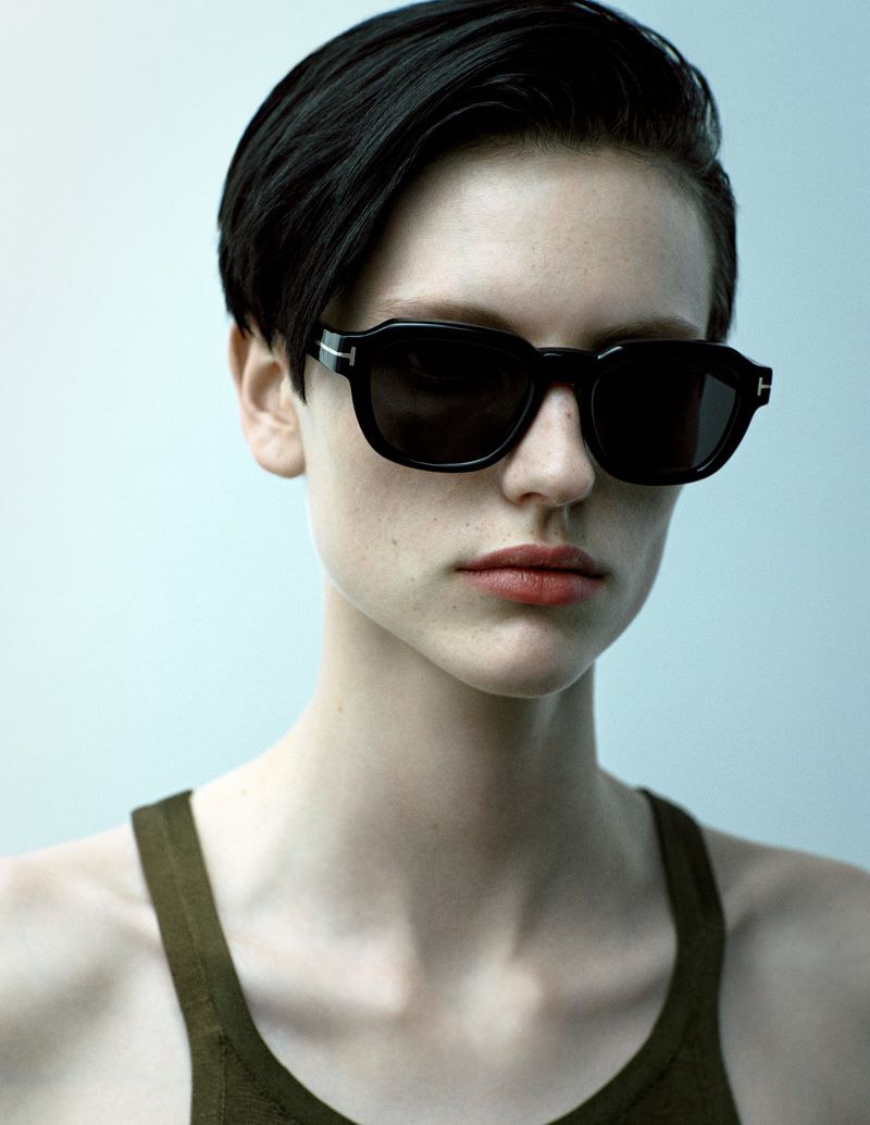 Close-up of an adult woman wearing dark sunglasses and an olive green tank top against a light blue background in portrait orientation.