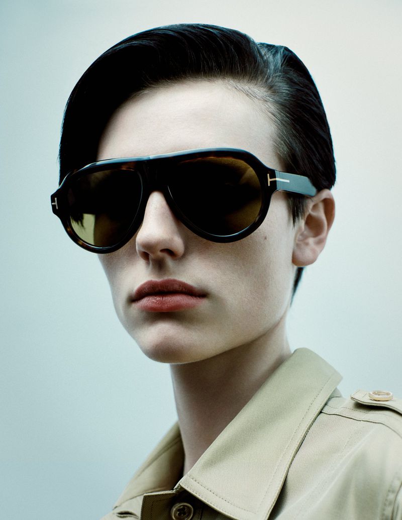 Woman with short hair wearing tortoise-shell aviator sunglasses and a beige trench coat stands against a light grey background.