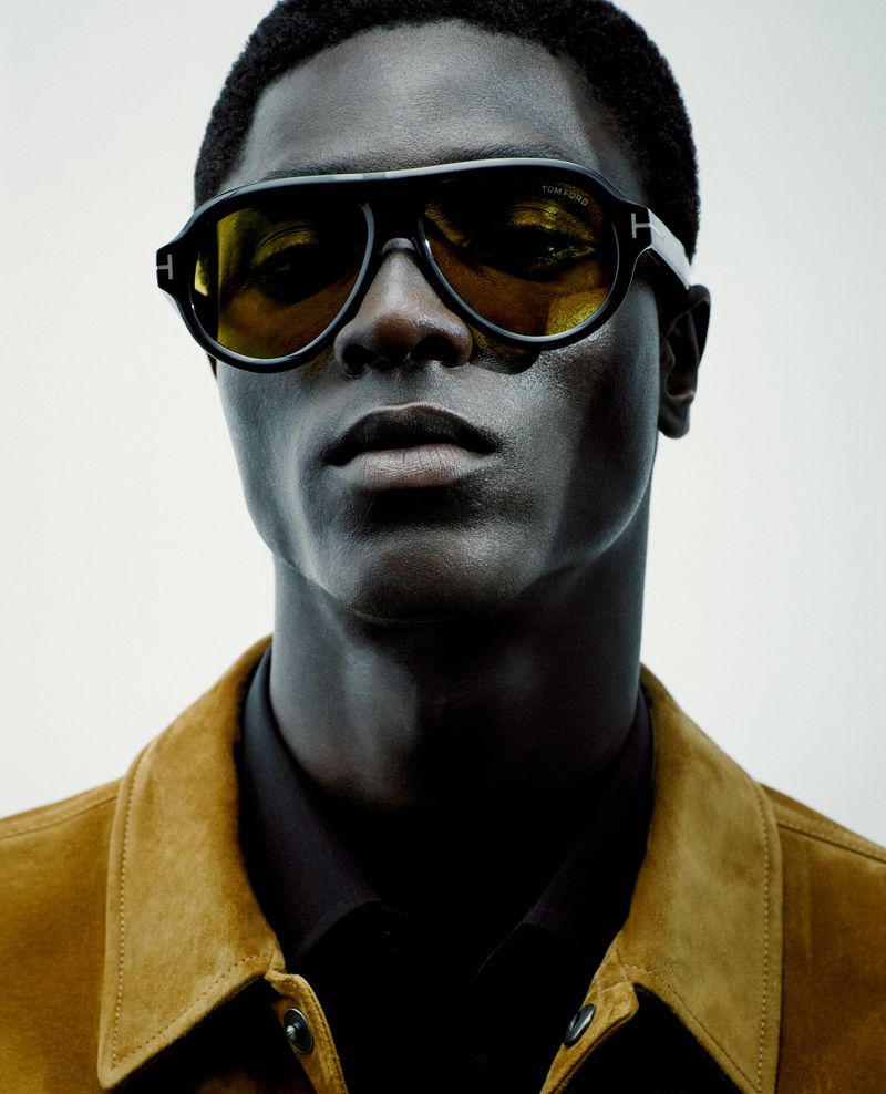 Close-up of a Black male fashion model wearing aviator sunglasses and a tan jacket against a neutral background in a studio setting.