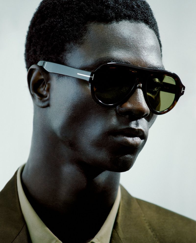 Close-up shot of a dark-skinned man wearing dark aviator sunglasses and a khaki shirt with an olive green suit jacket against a light gray background.