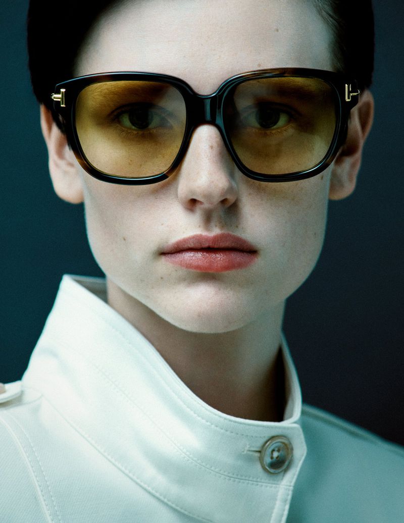 Close-up of an adult female model wearing tortoise shell sunglasses and a light jacket in a studio setting with dark gray backdrop.
