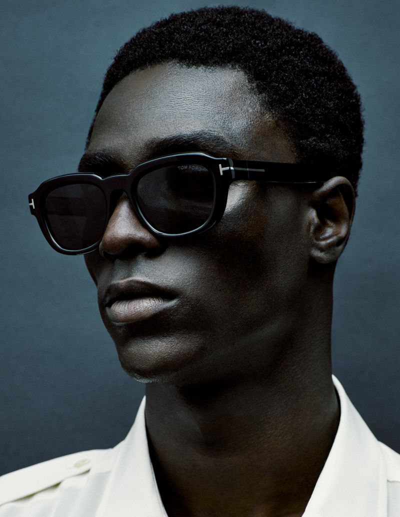 Close-up of a Black male model wearing black sunglasses and a light shirt against a dark blue background in a fashion portrait.