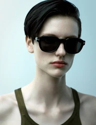 Model wearing dark sunglasses and an olive green tank top against a pale neutral background