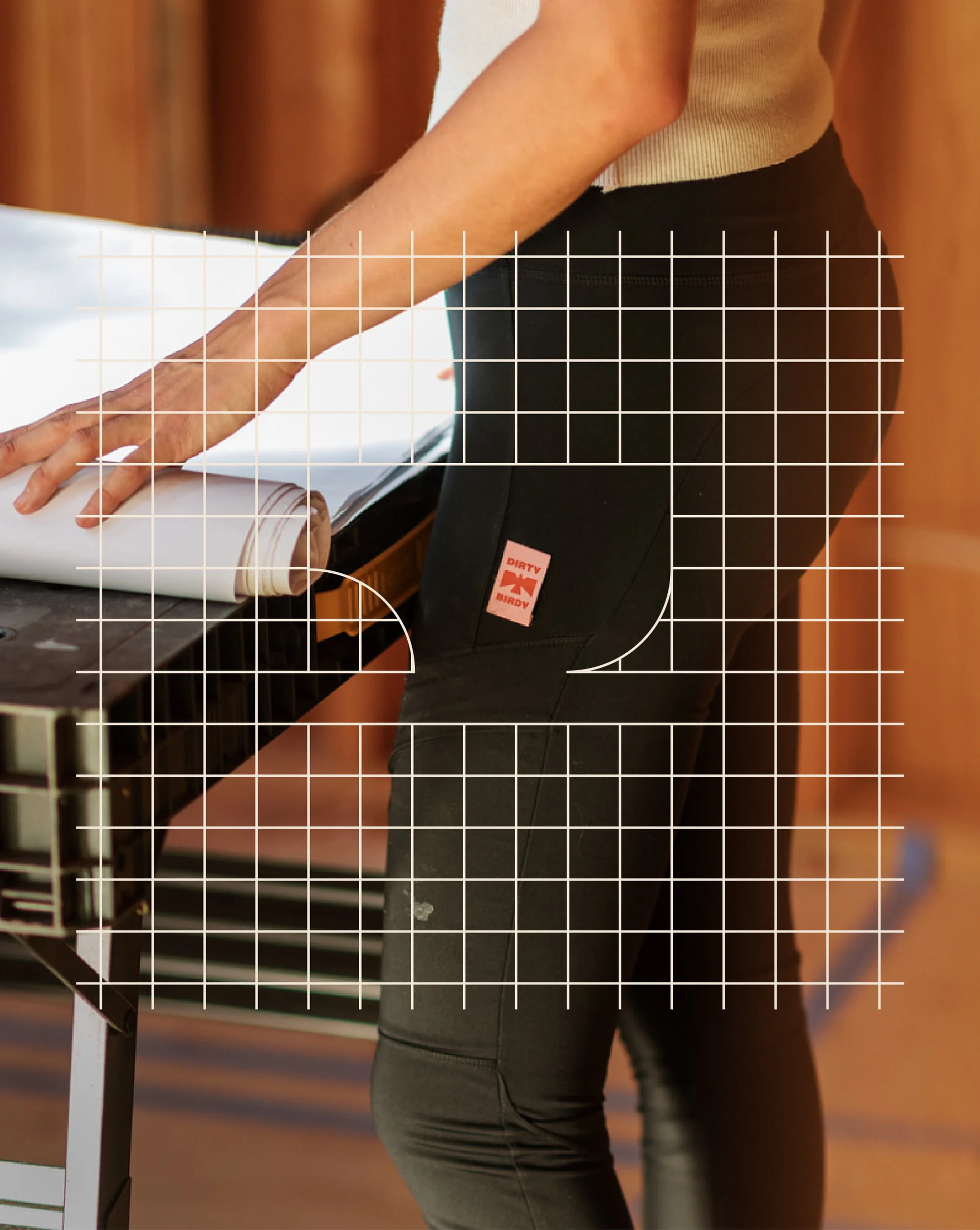 Woman in dark work pants rolls out a blueprint on a worktable inside an unfinished building, showcasing the garment's fit and style.