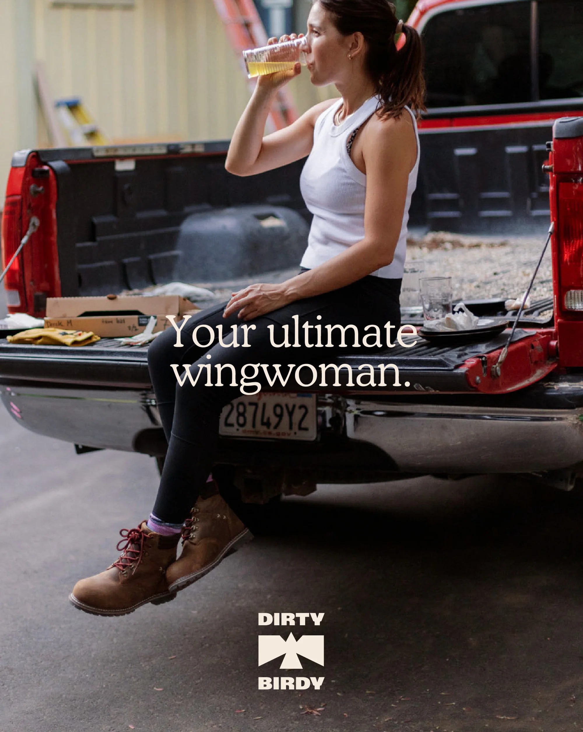 A woman in workout clothes drinks from a clear plastic cup, sitting on the back of a parked red pickup truck with construction in the background.