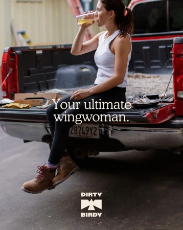 A woman in workout clothes drinks from a clear plastic cup, sitting on the back of a parked red pickup truck with construction in the background.