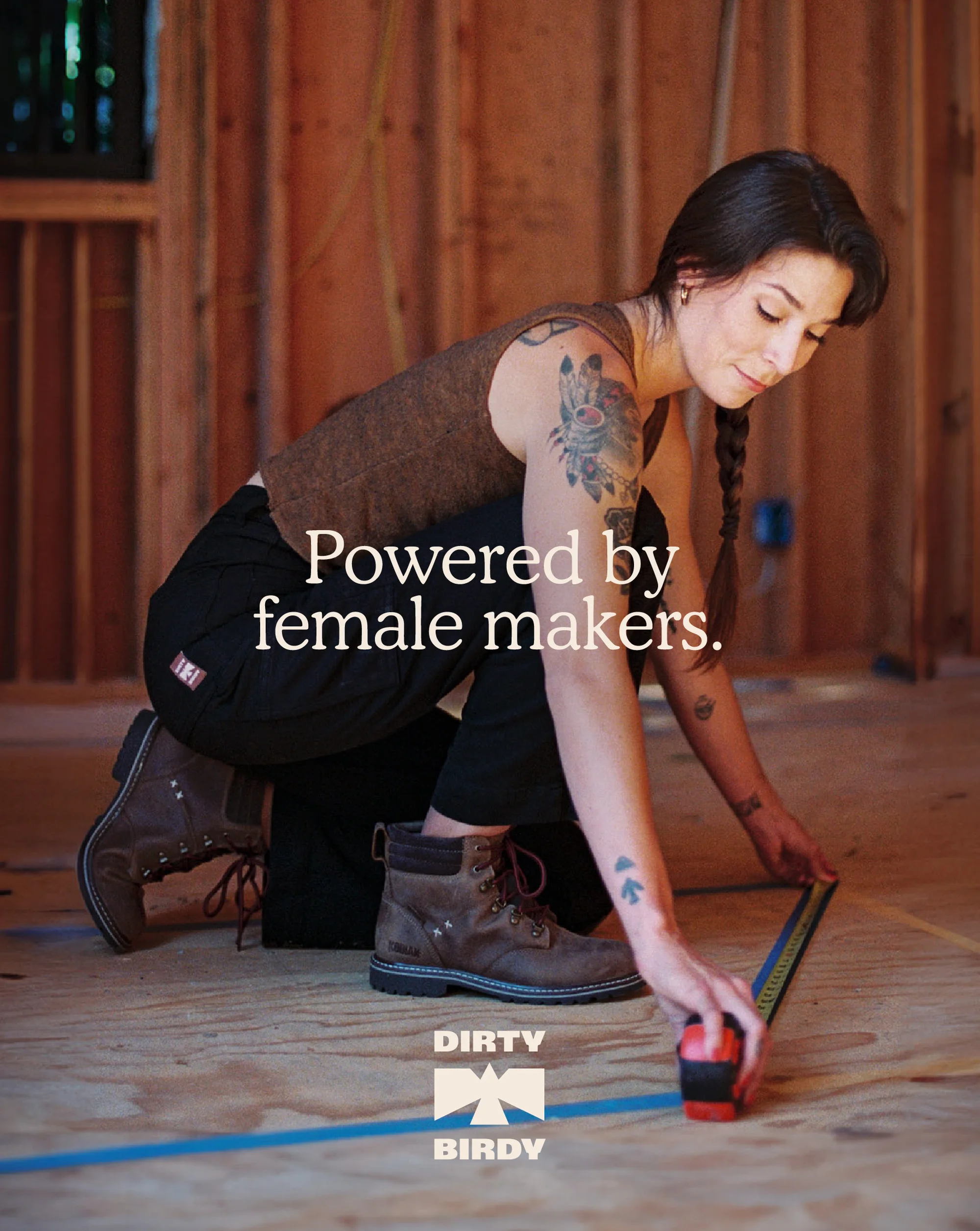 Brunette woman with arm tattoos measures wood planks in an unfinished home interior, wearing work pants, brown boots, and a brown tank top.