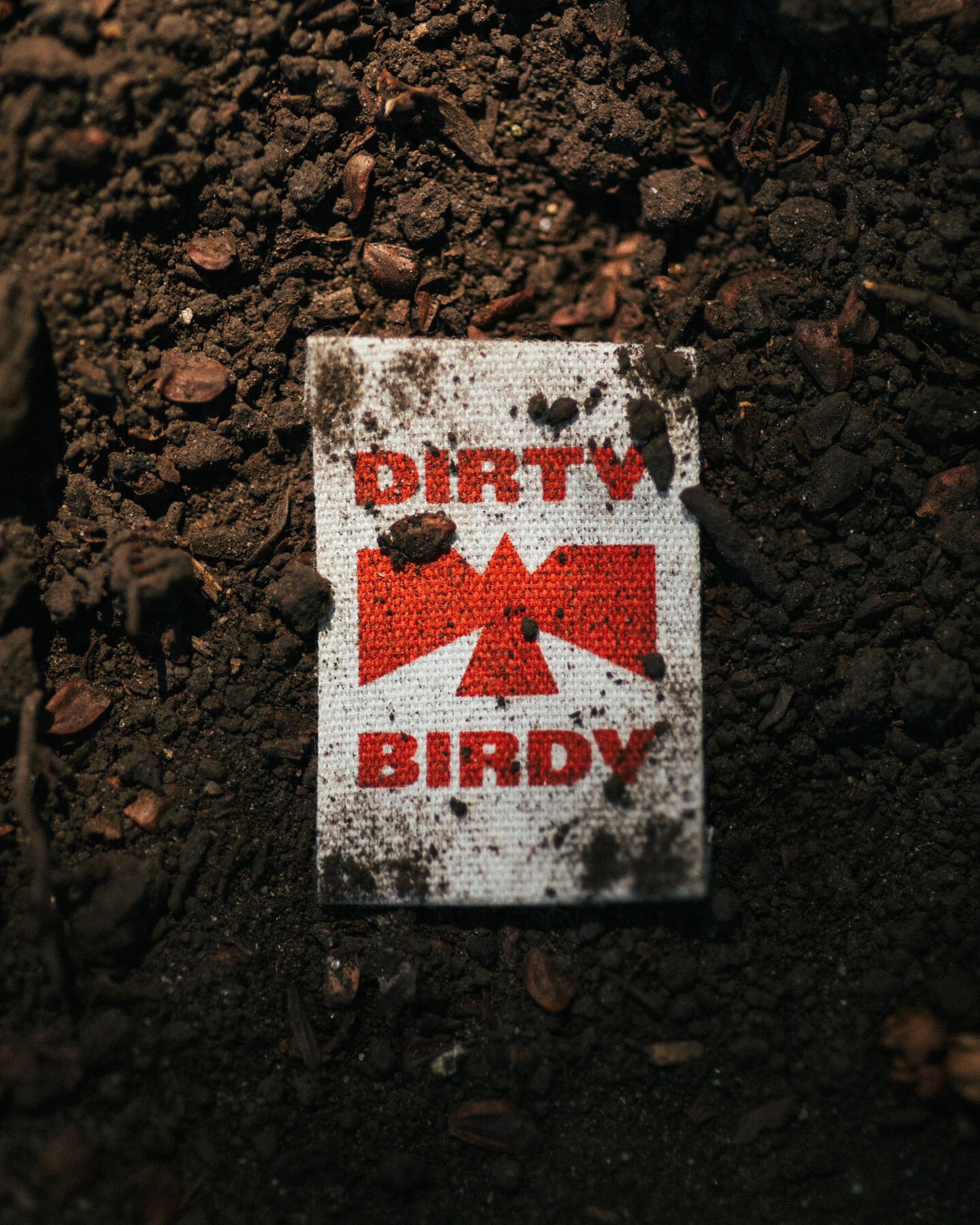 Dirty Birdy branded patch laying on dark brown soil, close-up shot.