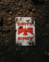 Close-up of a white woven clothing label with red Dirty Birdy typography and bird logo embedded in dark soil.