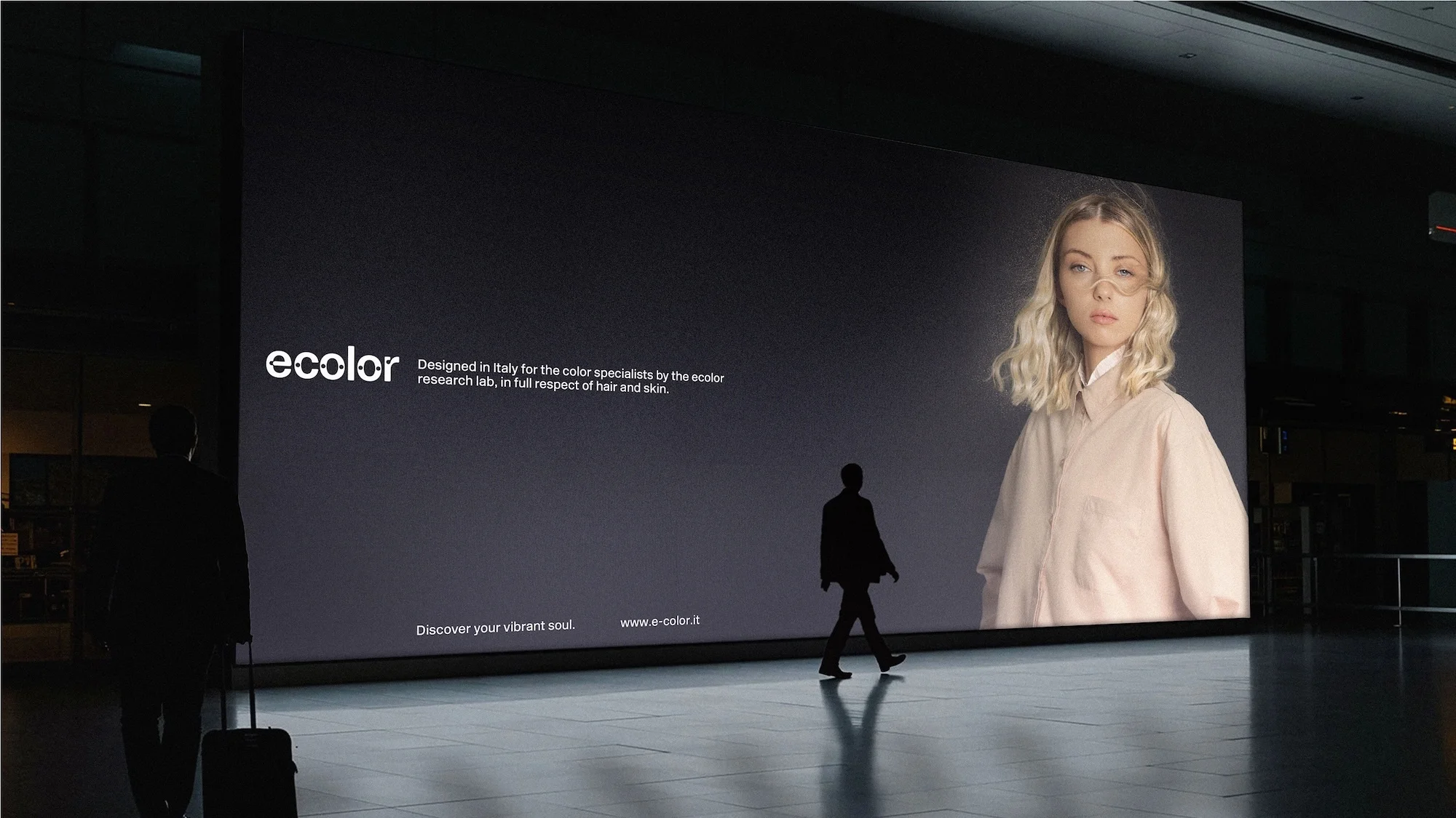 Billboard in an indoor space features a woman in a pink jacket, with a silhouette of a person walking in front of the display.