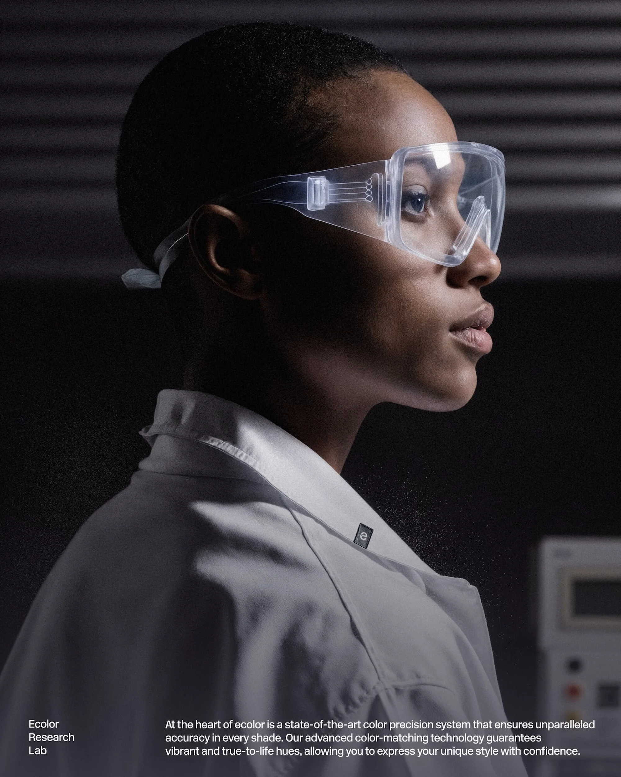Close-up of a woman in safety glasses and a lab coat, side view, in a research lab setting with scientific equipment in the background.