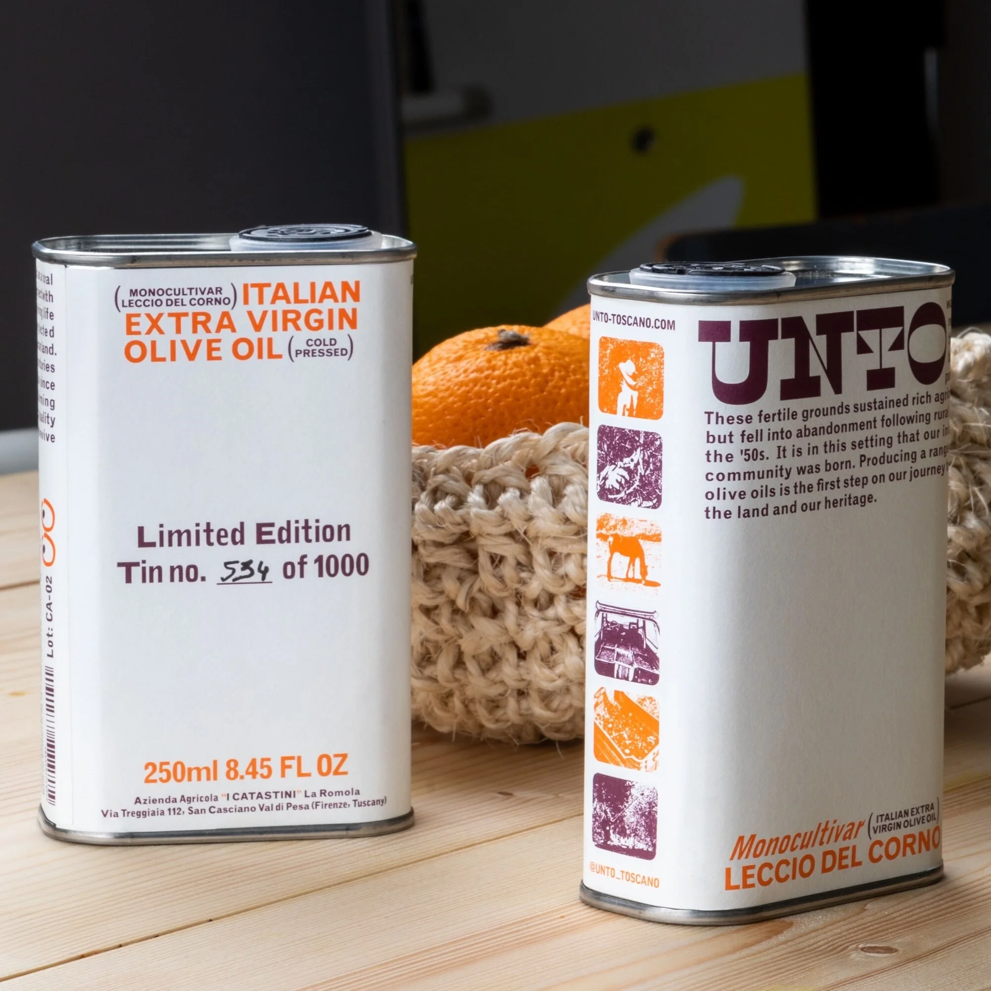 Two tins of Italian extra virgin olive oil sit on a wooden surface with oranges in a woven basket in the background.