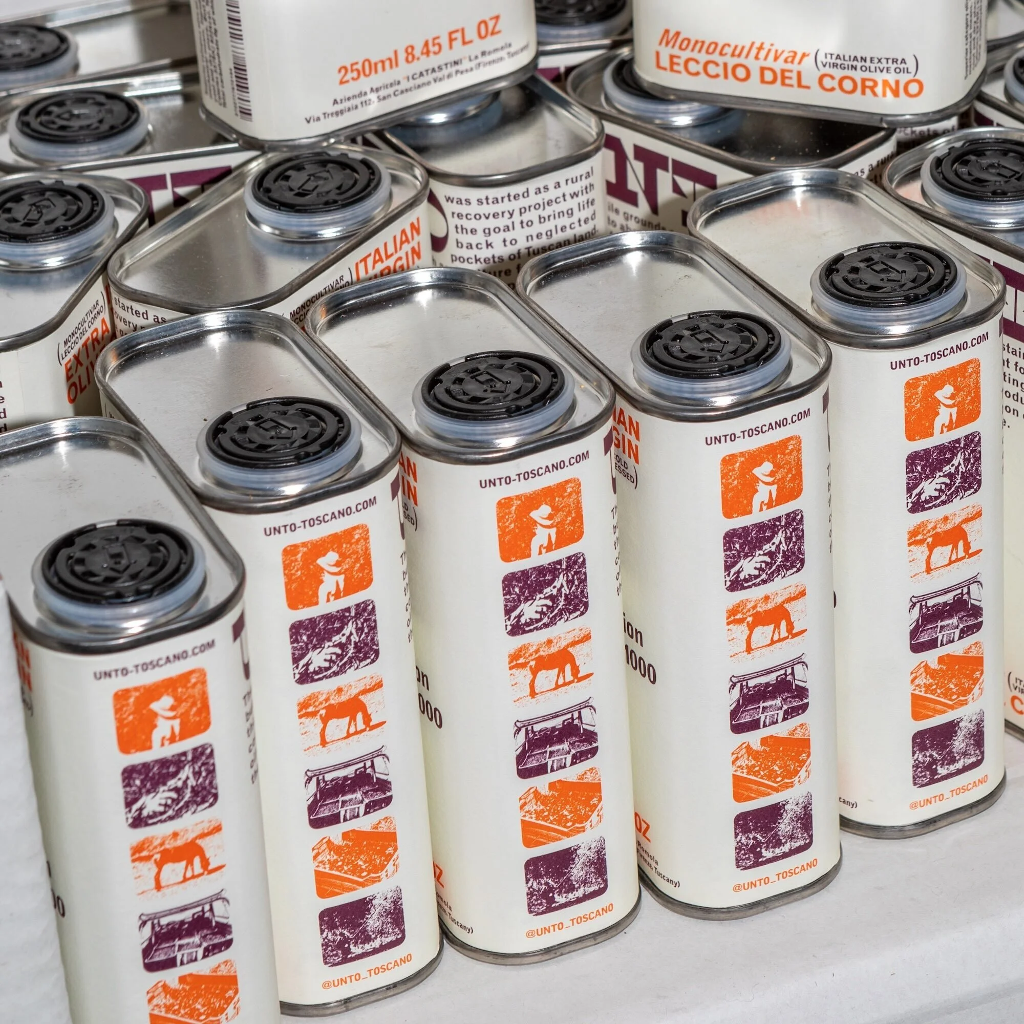 Multiple cans of Italian olive oil with white labels and graphic stampings in shades of orange and purple. The cans have black lids.