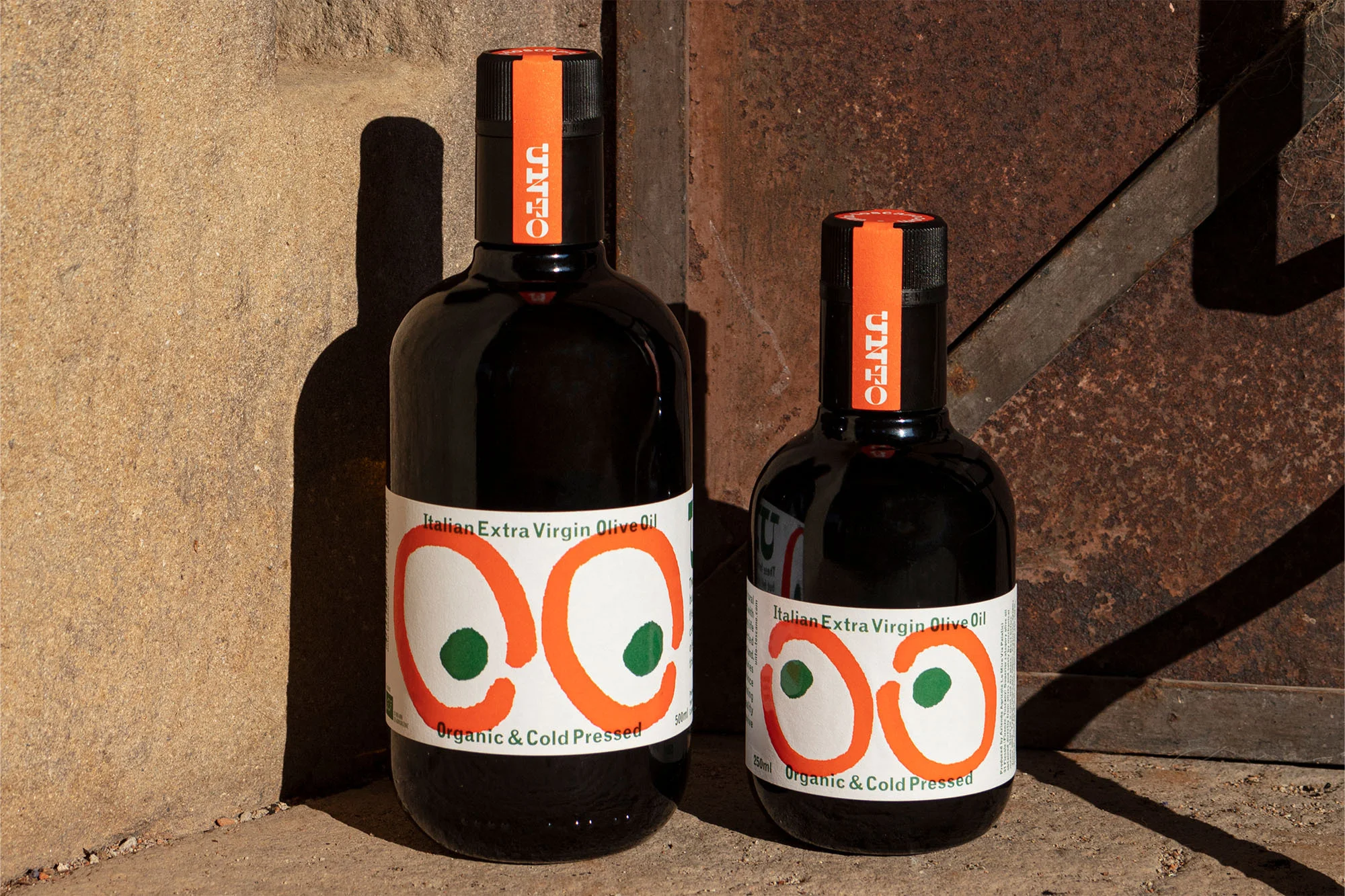 Two bottles of Italian Extra Virgin Olive Oil with orange and green logo on a white label, standing on a concrete surface.