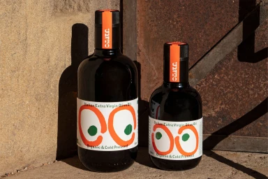 Two dark glass olive oil bottles with orange and green labels standing outdoors against a weathered metal wall