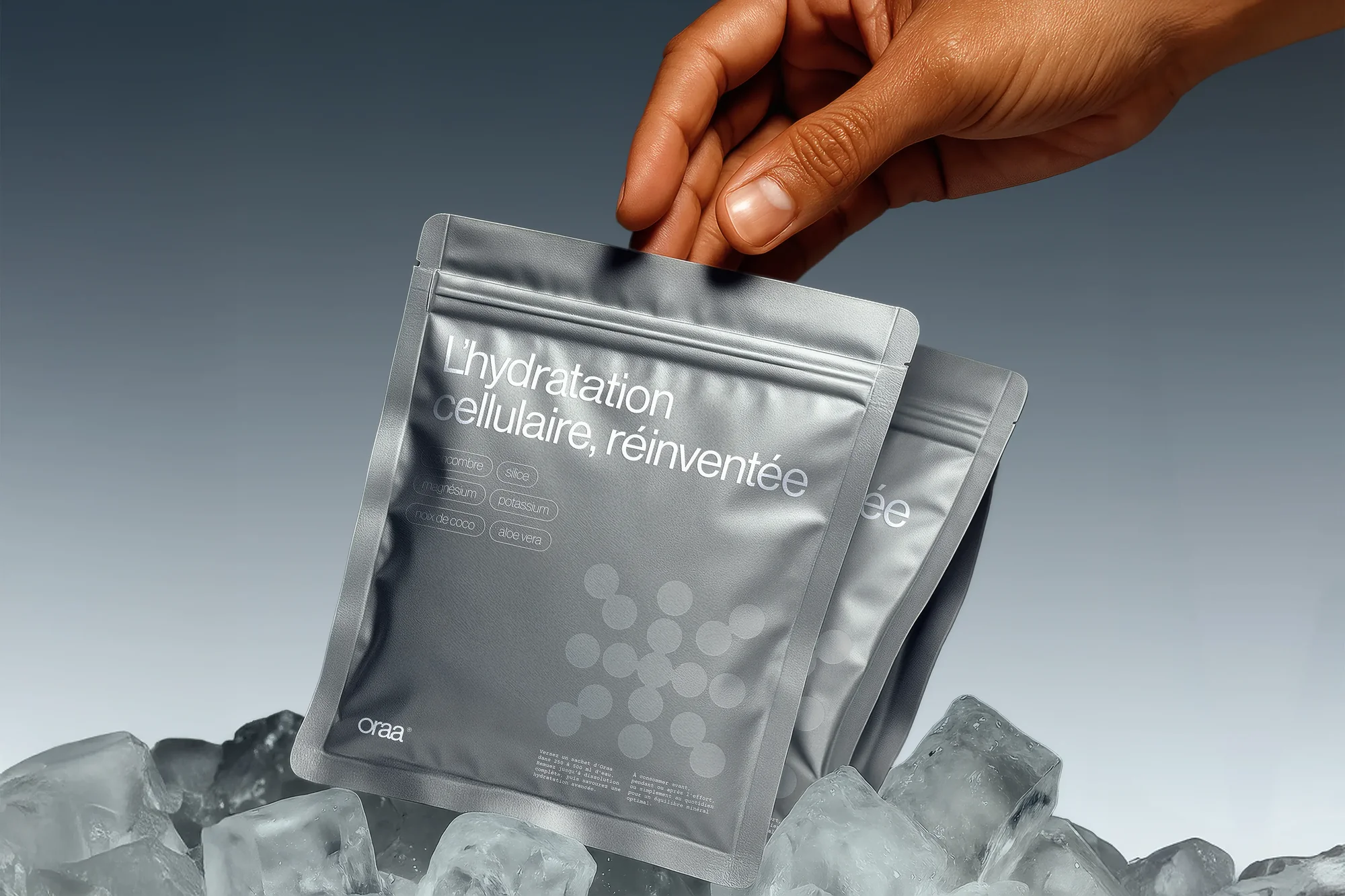 Hand reaching for hydration packets in ice. Silver pouches labeled 'L'Hydratation cellulaire, reinventee' on a grey background.