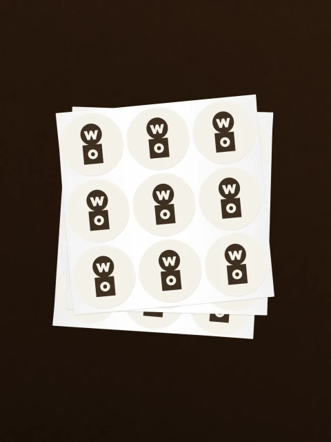 Sheet of round stickers with a 'WO' logo in black on an off-white background, stacked on a dark brown surface, eye-level, studio.