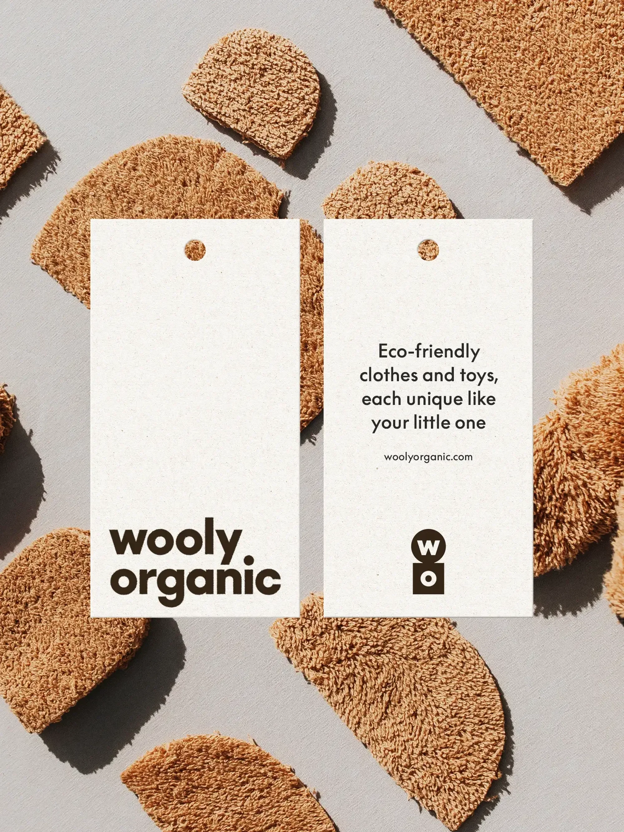 Two white product tags for Wooly Organic brand children's clothing surrounded by textured brown fabric samples on a gray surface.