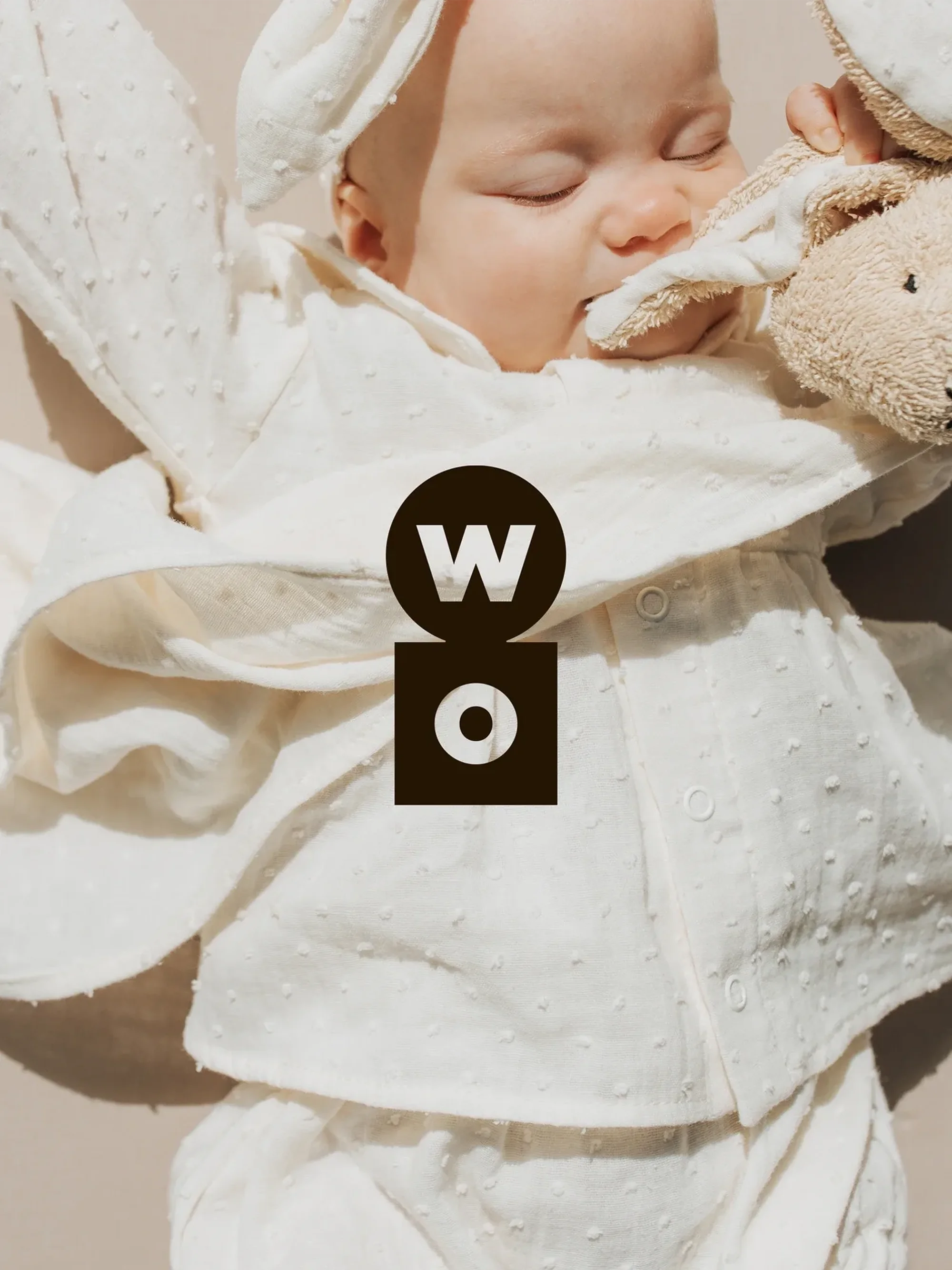 Infant lies on its back in white textured clothing holding a stuffed bunny in a neutral setting, promoting baby or infant clothing.