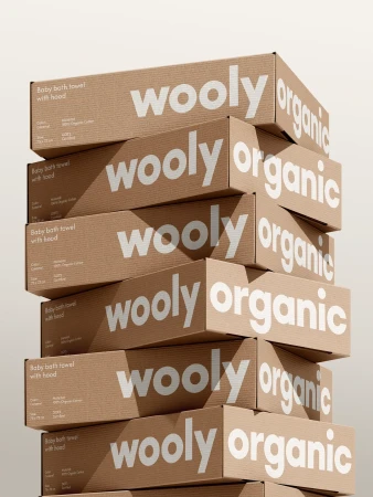 Stacked packaging boxes of Wooly Organic baby bath towels with hood. Tan cardboard boxes with white logos and product descriptions.