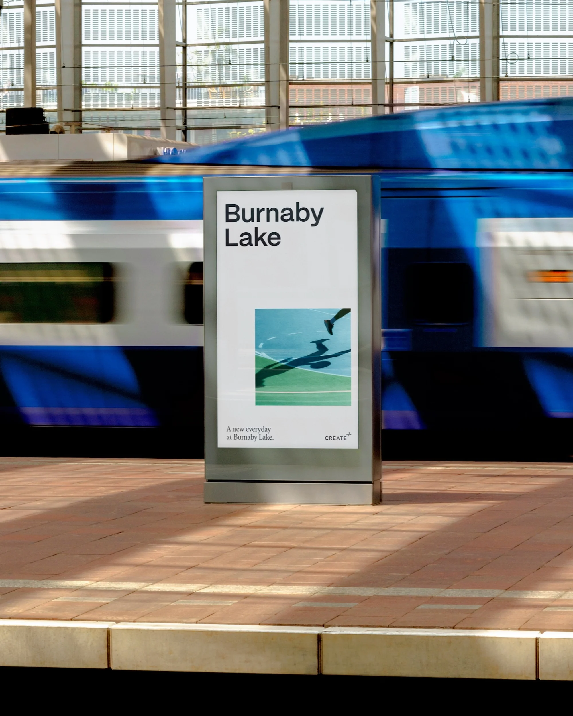 Motion blur of a blue train passing a transit advertisement for Burnaby Lake at a station with brick platform and windows.