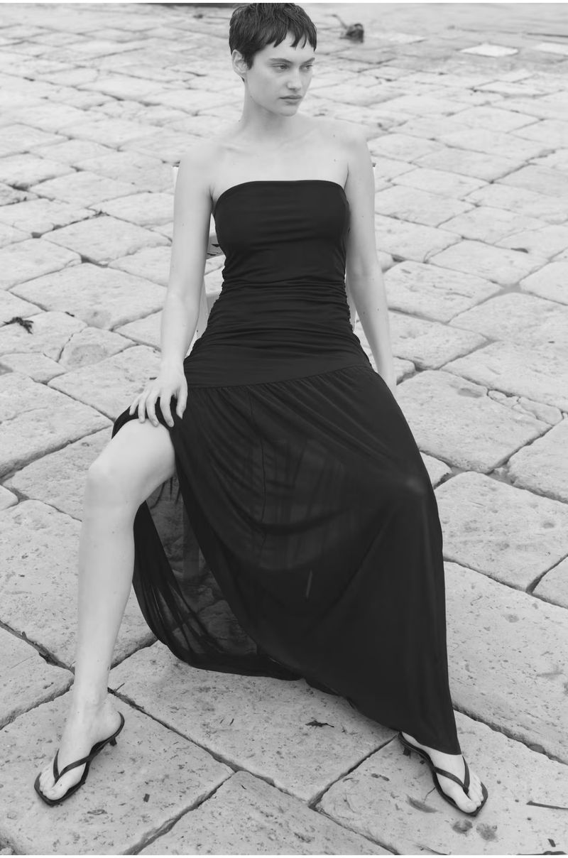 A woman sits outdoors on stone, wearing a strapless black dress and sandals. The image is in black and white with shallow depth of field.