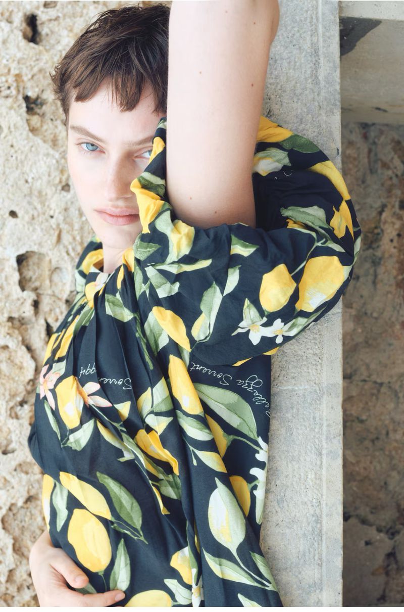 Portrait of a woman with short brown hair wearing a black top with lemon print leaning against a stone column with neutral expression.