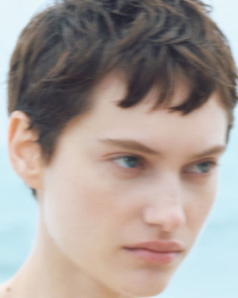 Close-up portrait of a young adult female with short dark hair and fair skin against a blurred background of the sky and water.
