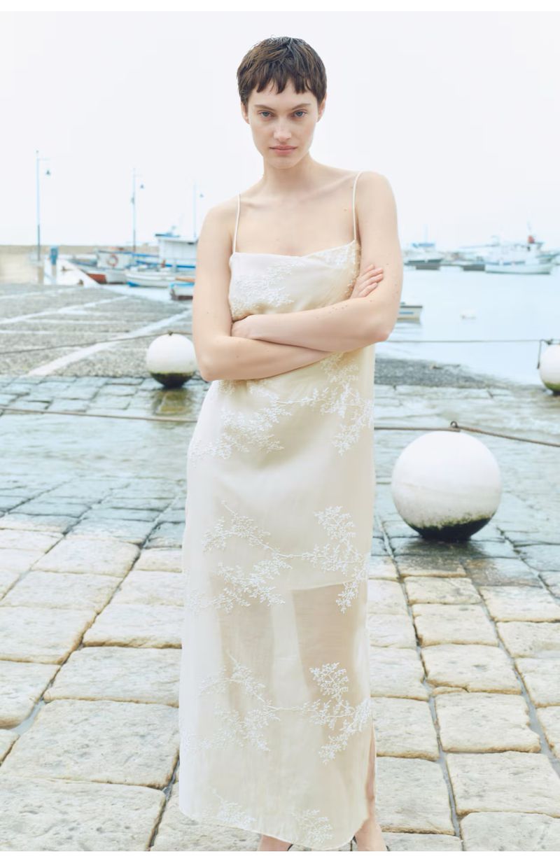A woman stands on a stone dock in a cream-colored slip dress with floral embroidery, posing with her arms crossed in a fashion shot.
