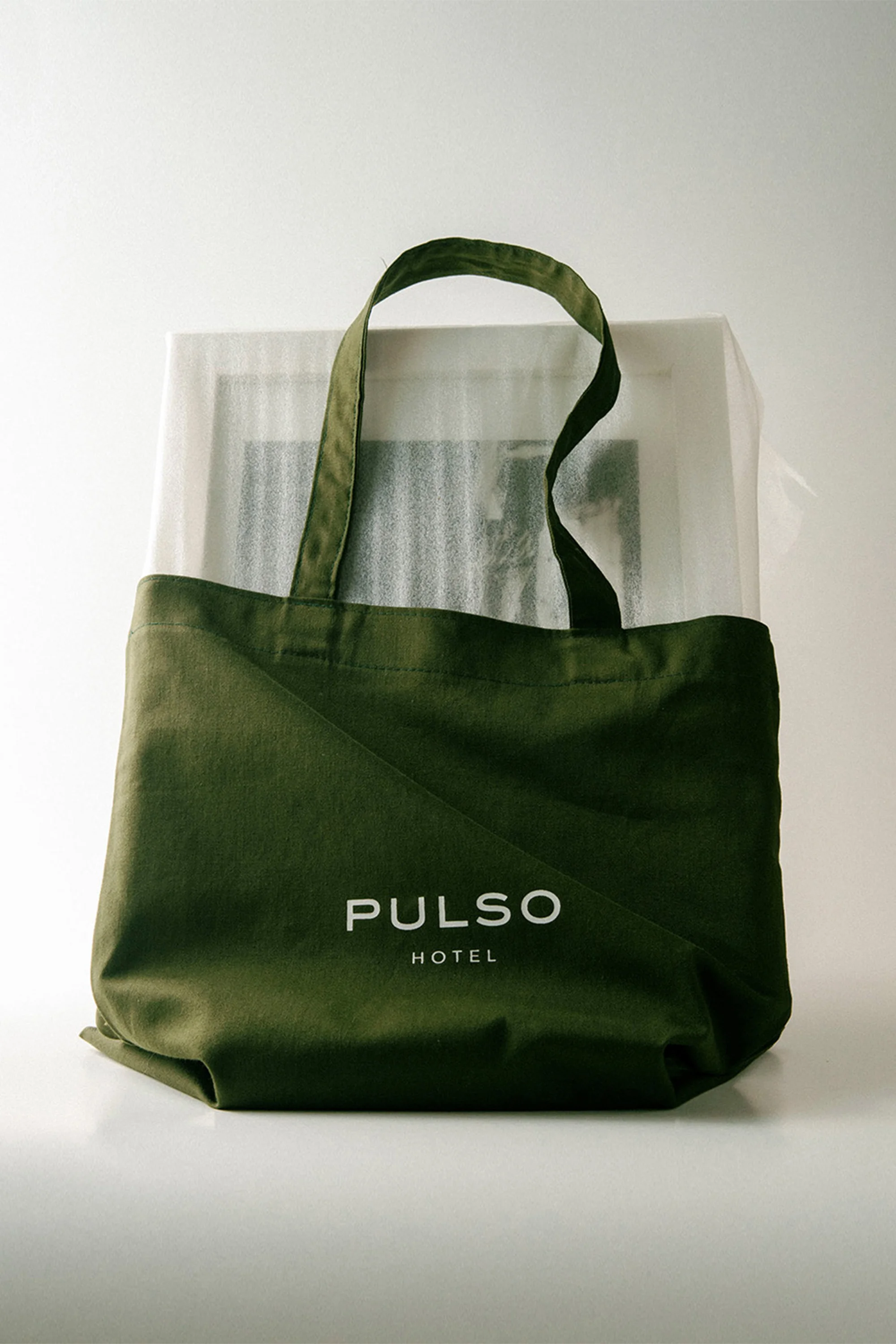 Olive-green tote bag with 'Pulso Hotel' branding, containing a wrapped framed object, against a bright white background.