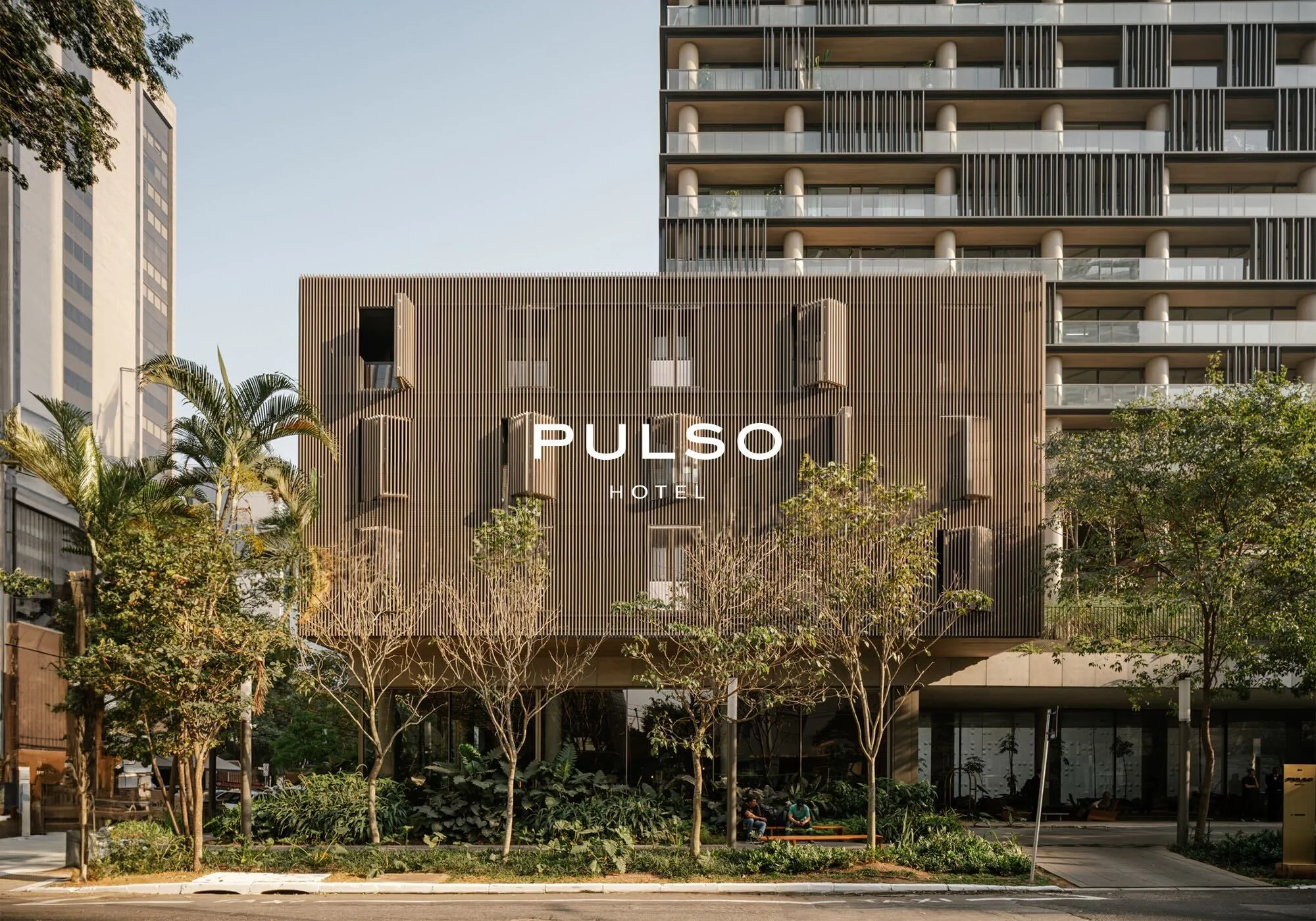 Exterior view of PULSO Hotel showcasing a modern slatted wood facade and illuminated signage amid mature trees and street landscaping.