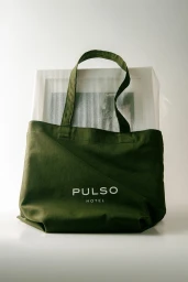 Dark green canvas tote bag featuring the Pulso Hotel logo, placed in front of a white framed piece against a neutral backdrop