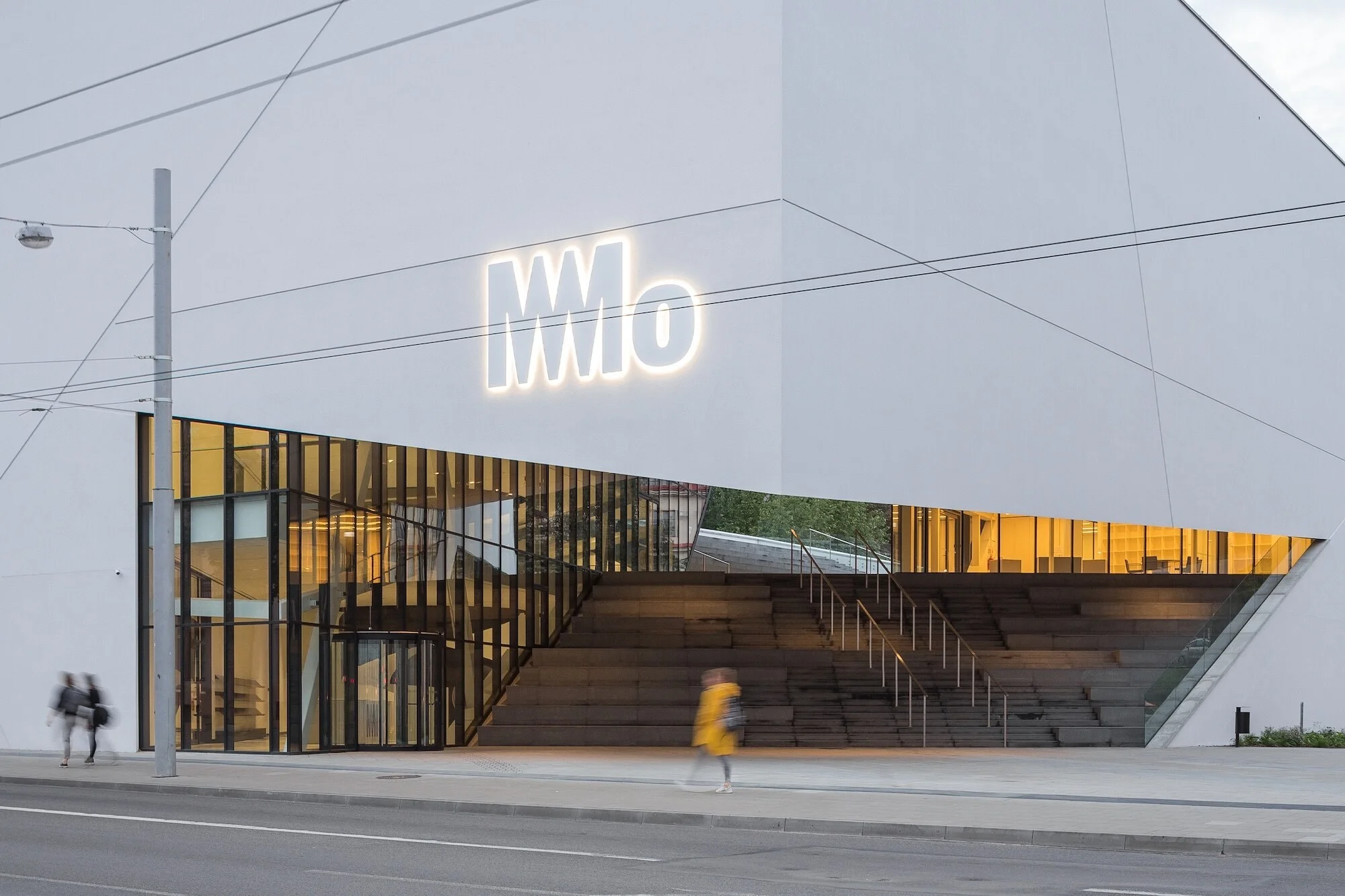 Modern architecture showcases a sleek white building with a prominent MMo sign, complemented by steps and blurred figures outside.