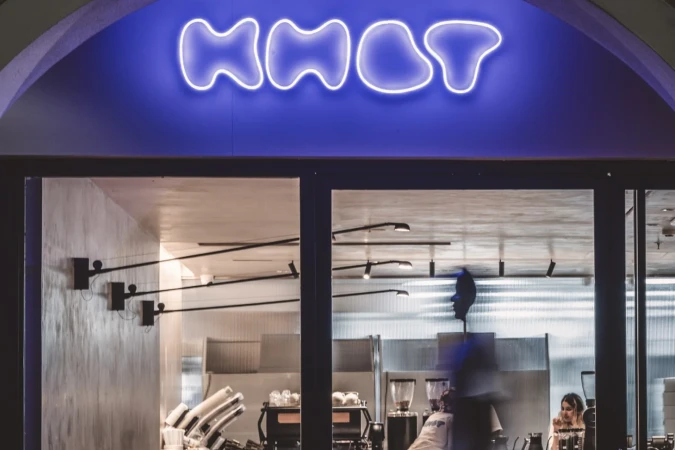 Exterior view of a cafe with a neon sign, interior lighting, and people visible through the window, creating an inviting atmosphere.