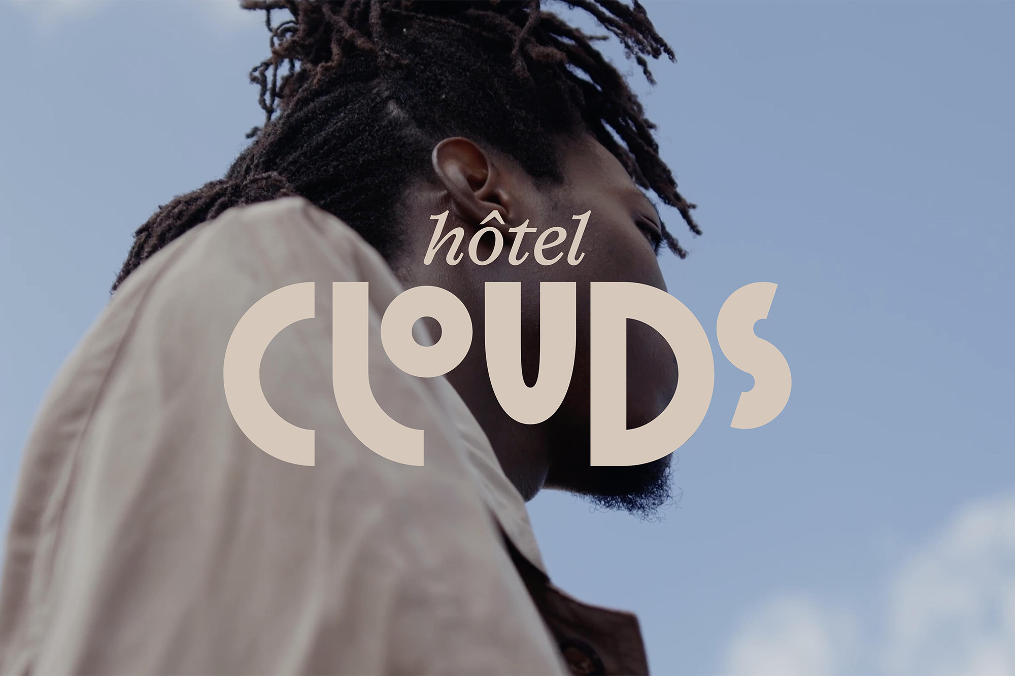 Portrait of a man with dreadlocks, wearing a tan jacket, against a blue sky with clouds, with the text 'Hotel Clouds' overlaid.