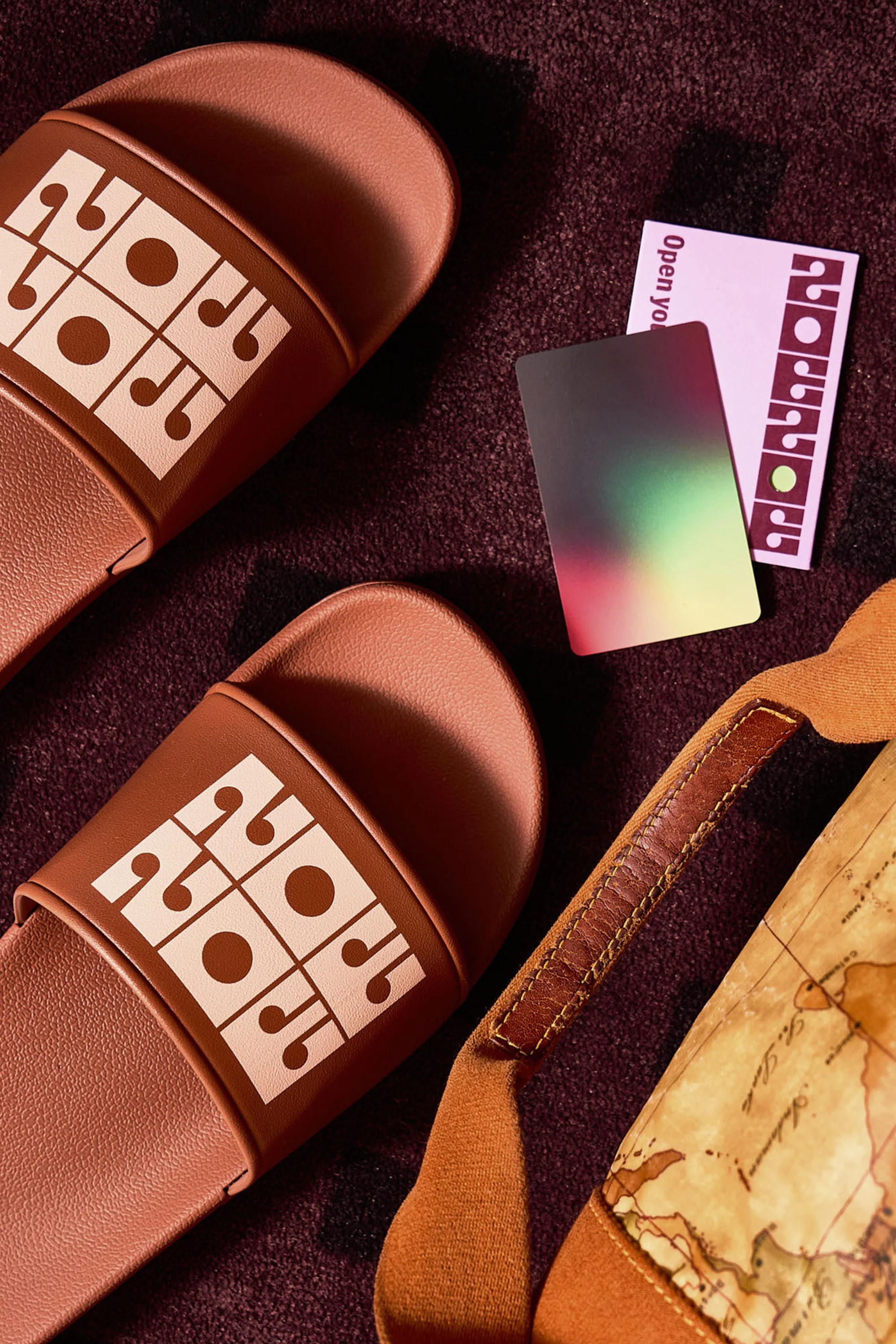 Flat lay studio shot of two brown slides with a geometric design, a card, a blurred card, and a brown travel bag on a textured surface.
